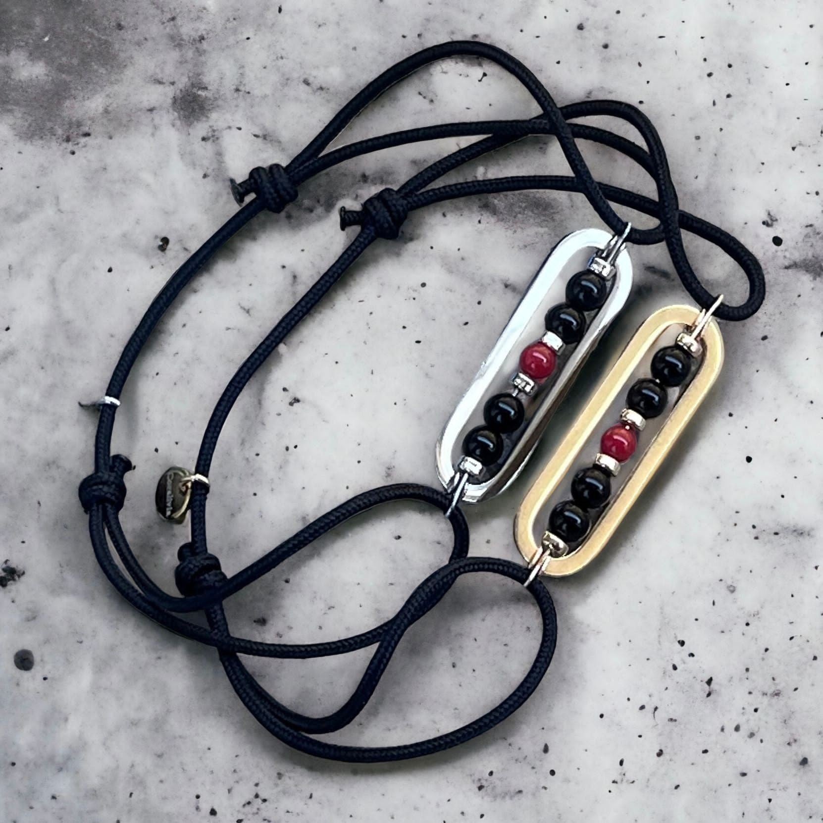 Bracelet homme Lesio fait main en Corse avec perles d’Onyx noir, perle centrale en corail rouge de Méditerranée, rectangles en acier inoxydable doré et argenté, cordons noirs ajustables avec nœuds coulissants
